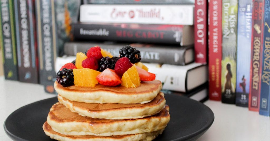Stack of pancakes topped with fresh berries and oranges against a backdrop of books.